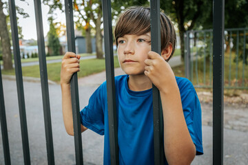 Obraz premium A young boy in a blue shirt grips metal bars while standing in a park, looking directly at the camera. The background features green trees and a fence
