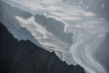Dramatic alpine view to large glacier among rocks and ridges in gray low clouds in harsh weather. Dark atmospheric mountain landscape with long big glacier tongue in rain. Ice and rockies close up.