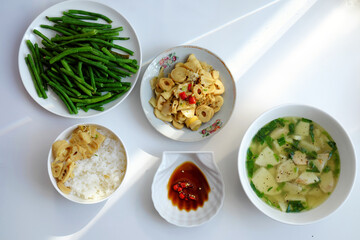 Vietnamese meal with steamed rice, tofu and vegetable soup, stir-fried green beans, bamboo shoots, and chili sauce on a white table, illuminated by natural light from a window.