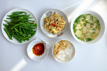 Vietnamese meal with steamed rice, tofu and vegetable soup, stir-fried green beans, bamboo shoots, and chili sauce on a white table, illuminated by natural light from a window.