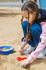 Pretty child girl playing in sand on outdoor playground. Beautiful baby in jeans trousers having fun on sunny warm summer day. Child with colorful sand toys. Healthy active baby outdoors plays games