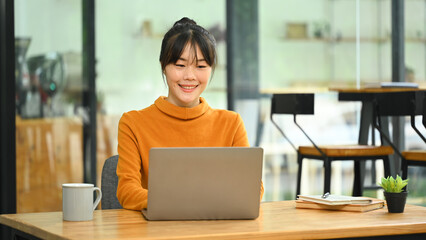 Pretty young Asian woman in yellow sweater using laptop in the cafe