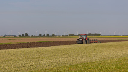 a red tractor is in the process oh plowing. In the distance are wind turbines. A natural setting with copy space in the picture