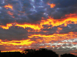 a breathtaking sunset with a dramatic sky filled with thick, textured clouds. The clouds, predominantly dark and foreboding, are illuminated by a vibrant orange and red glow, creating a stark contrast