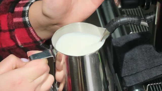 Barista froths milk into steel jug with espresso machine. Person holds pitcher as hand guides steam wand submerged in liquid slow motion