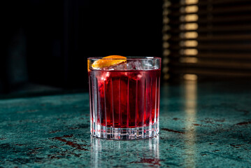 A glass of red cocktail with an orange slice garnish on a textured table, creating a refreshing and moody atmosphere. Perfect for summer drink concepts, bar menus, or lifestyle imagery.