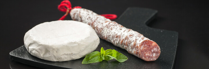Spanish fuet sausages cut in slices , cheese Brie and basil on a black cutting board. Black background