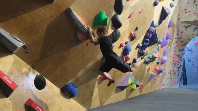Teenager climbs up rock face gripping synthetic stones. Fitness enthusiast tackles indoor training wall for alpinists slow motion