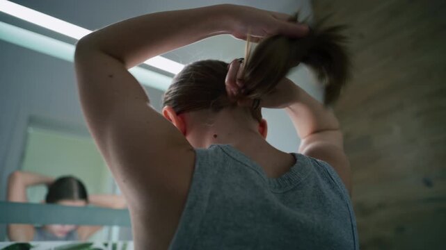 Pretty teenager stands in front of sink and ties her hair in ponytail before going to bed. Attractive young girl looks at the mirror in bathroom. Evening bath routine. Concept of lifestyle. Close up.