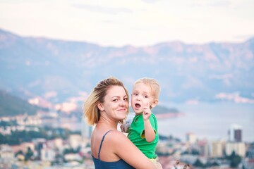 Mother and Son's Excited Discovery on Coastal Mountain Vacation. Blonde woman holds enthusiastic boy overlooking picturesque coastal town and mountains. Captures family travel joy and child's wonder