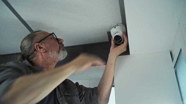 Professional male installer in uniform screws security camera to the ceiling in office room using tool and adjusts the angle. Monitoring and tracking system. Surveillance system, CCTV and privacy.