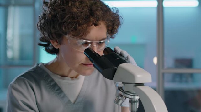 Medium close-up shot of young curly-haired Caucasian female scientist in protective goggles checking specimen glass, putting under microscope for studying and adjusting focus