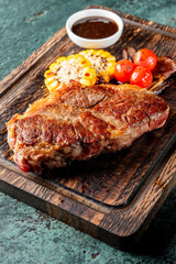 Grilled steak on a wooden board with corn, cherry tomatoes, and a bowl of sauce. The rustic presentation highlights the juicy, perfectly cooked meat and fresh vegetables, ideal for food photography.