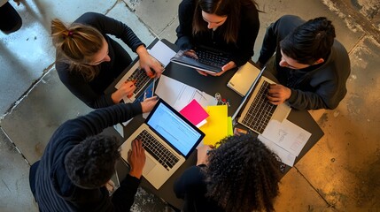 A diverse group of young adults work together around a table, using laptops and tablets.
