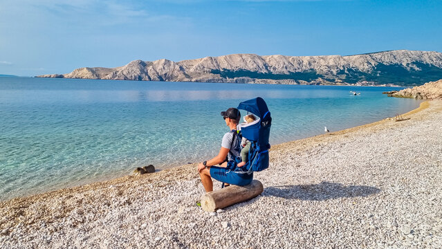 Man with baby carrier backpack in serene coastal scene on pristine pebble beach Vrzenica, Baska, Krk Otok, Primorje-Gorski Kotar, Croatia. Crystal-clear turquoise waters of Adriatic Sea. Hiking trail