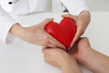 Obraz premium Doctor giving red heart to patient at white table in clinic, closeup