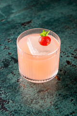 A pink cocktail in a short glass, garnished with a green leaf and red berry, rests on a textured...