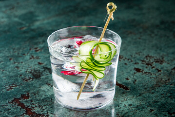 A clear glass with ice cubes holds a refreshing drink garnished with cucumber and red berries. This...
