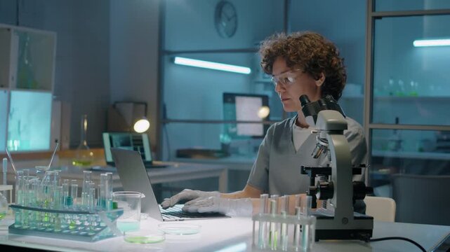 Medium tilt shot of young Caucasian female biochemist in uniform, goggles, gloves adjusting focus, observing microbes under microscope, diagnosing abnormalities and typing report on laptop
