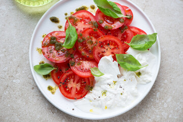 Red tomatoes with basil oil, fresh basil and burrata cheese served on a white plate, horizontal shot on a beige stone surface, elevated view
