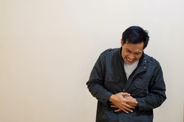 A Southeast Asian man wear brown jacket, in pain holding his hurting stomach isolated on the cream...