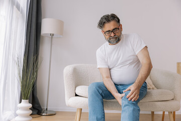 Home, knee pain and senior man with arthritis, osteoporosis and fibromyalgia on sofa. Elderly male...