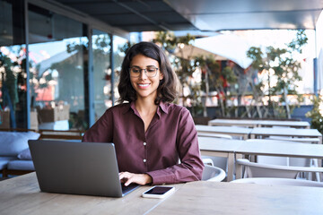 Young latin or arabian freelance female student worker using laptop pc sitting outdoors at cafe office. Portrait of middle eastern indian business woman working remotely on finance project. Copy space