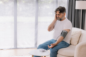 Handsome grey-haired senior man in casual outfit sitting on couch at home, checking blood pressure on his own. Pensioner using modern tonometer, experiencing hypertension, copy space