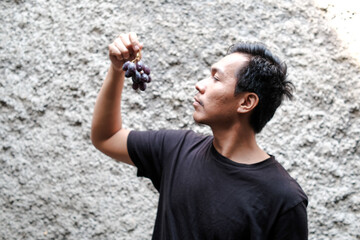 Fototapeta premium A Southeast Asian man eating grape fruits in outdoor with grey wall, vegetarian, diet food, sweet fruits.