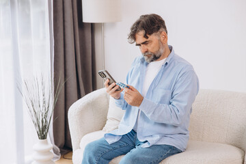 An elderly man is sitting alone on the couch a patient is wearing glasses reading instructions on a...