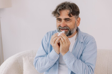 Senior man sneezing into a tissue, experiencing discomfort from cold or allergies, seasonal...