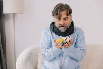 Senior gray bearded excited man in scarf having acute viral infection holding many anti-inflammatory drugs in hands in living room at home