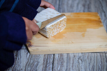 Close up Female Hand Slicing Cut Tempeh or Tempe on Wooden Chopping Board Using Knife. Tempe is Fermented Soy Product Originally from Indonesia.