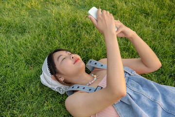 Happy woman lying down on the grass enjoying nature. Lying down and playing with your cell phone in an outdoor park, smiling brightly, immersing yourself in the nature around you