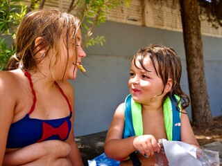 Close-up portrait of two young caucasian siblings laughing and sharing a snack outside on a sunny summer day. Girl in swimsuit and boy in life jacket.