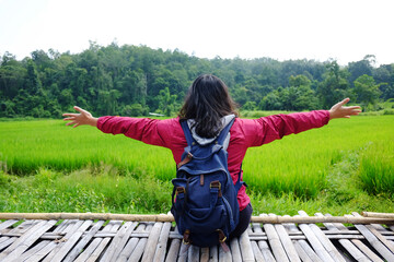 Happiness of Asian woman sitting and extend arms for freedom on woven wooden bridge for walking across agriculture field and greenery paddy rice terrace on hill in Thailand
