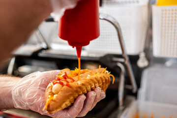 close-up of a hand in transparent glove squeezing ketchup from a bottle onto a hot dog topped with grated cheese. The background is blurred, emphasizing the action. Ideal for food and culinary themes.