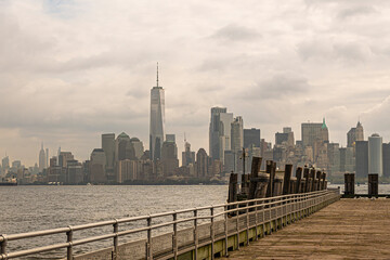 Fototapeta premium New York, NY, USA - August 4, 2023: Docking pier at Miss Liberty Island. Lower Manhattan in back under gray cloudscape