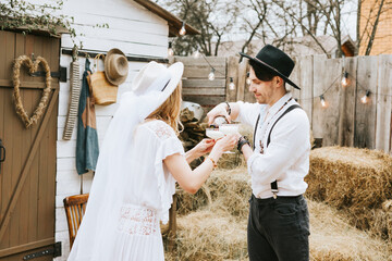 wedding boho style ceremony young couple newlyweds bride in white dress with veil and groom in hat and jeans with suspenders hugging and having fun at barn of country house, shot in motion
