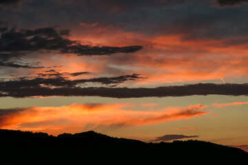 Dramatic sky in Jennersdorf, Burgenland, Austria. Thick swirling clouds painted in hues of gray, orange, pink. Sunset of warm glow creating breathtaking spectacle. Silhouette of rolling hill landscape