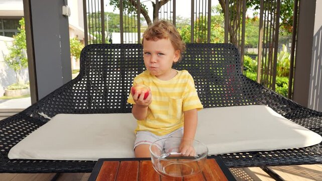 Adorable little boy enjoys flat peach, taking small bites and chewing sweet, slightly firm fruit. Sitting in modern outdoor gazebo on sunny morning, cheerful toddler savors his breakfast snack.