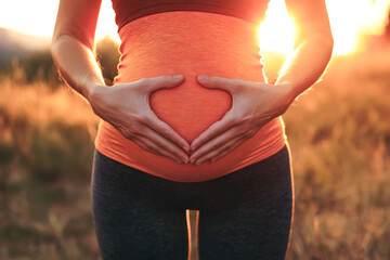 Pregnant woman with heart shaped hands around  belly wearing sport clothes with natural outdoor background.