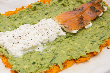 Close-up of a delicious layered dish featuring a base of grated carrots, a middle layer of creamy guacamole, and a topping of white cheese and smoked salmon with herbs on baking paper.