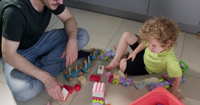 Father plays and toddler boy play with constructor set blocks sitting on floor. Careful parent teaches child to build simple items at home