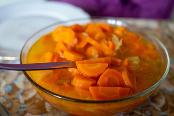 Bowl of cooked carrots in a savory broth with a serving spoon on a floral tablecloth.