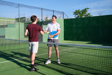 Two male paddle tennis players shaking hands through the net after a match on a sunny day