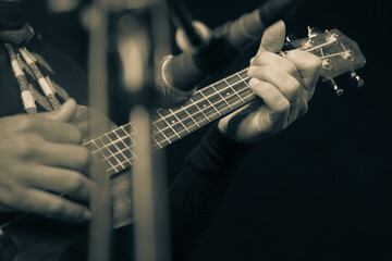 Close-up of a musician's hands playing a ukulele in black and white. The image captures the focus and skill of the performance, with a microphone in the background adding depth to the scene........