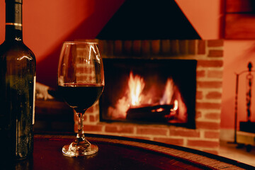 A glass of red wine and a bottle beside it are in sharp focus, while a lit fireplace casts a warm glow in the cozy winter background. The scene exudes comfort and relaxation.