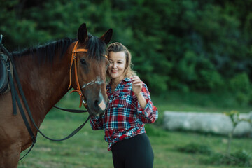 Obraz premium Happy blonde with horse in forest. Woman and a horse walking through the field during the day. Dressed in a plaid shirt and black leggings.