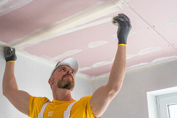 The worker make a plasterboard ceiling. He does taping plaster drywall ceiling joints. He is ...